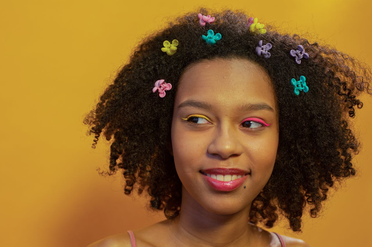 Close-up portrait of a smiling woman with afro hair, colorful hair clips, and eye makeup against a yellow background.