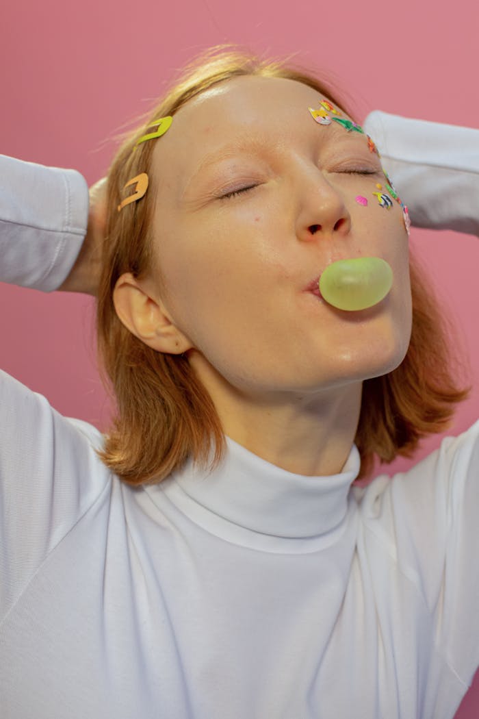 Young lady in casual clothes and colorful animal stickers on face with hairpins in hair blowing gum bubble with closed eyes and hand behind head on pink background in studio